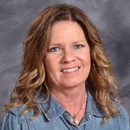 Smiling woman with wavy hair wearing a denim shirt and pearl necklace